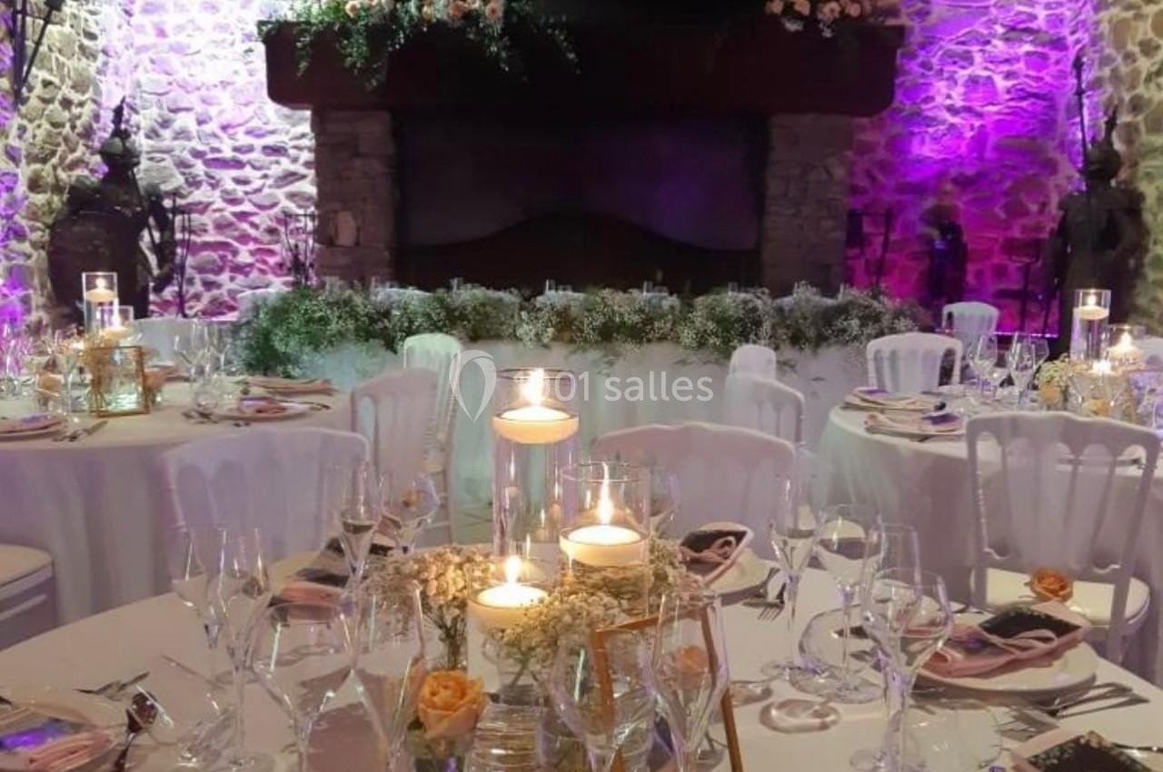 Salle de réception décorée pour un mariage, avec tables dressées, bougies et éclairage violet sur des murs en pierre.