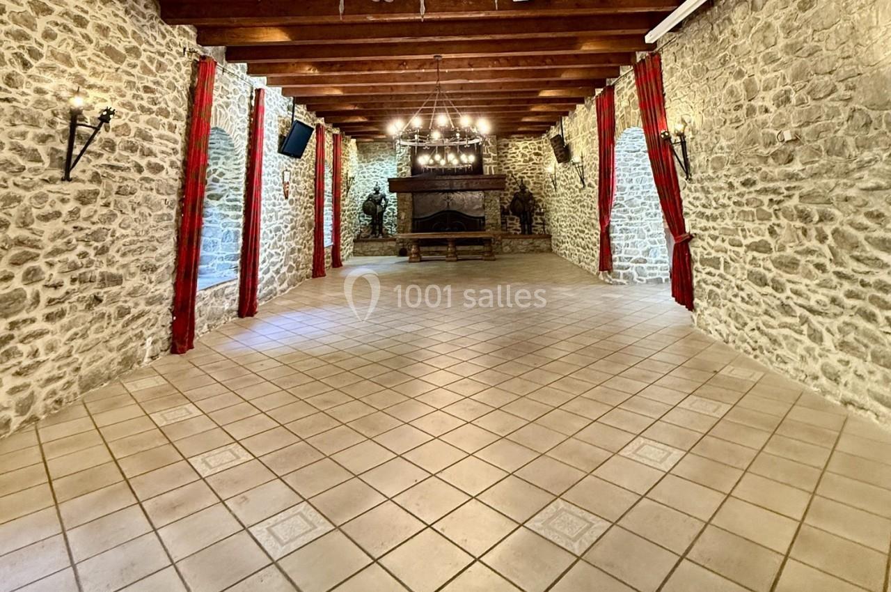 Salle en pierre avec poutres apparentes, sol carrelé, cheminée centrale et rideaux rouges encadrant les murs.