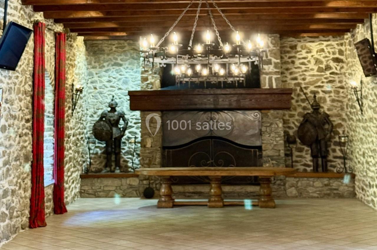 Salle en pierre avec une cheminée imposante, un lustre en fer forgé, des statues de chevaliers et des rideaux rouges.