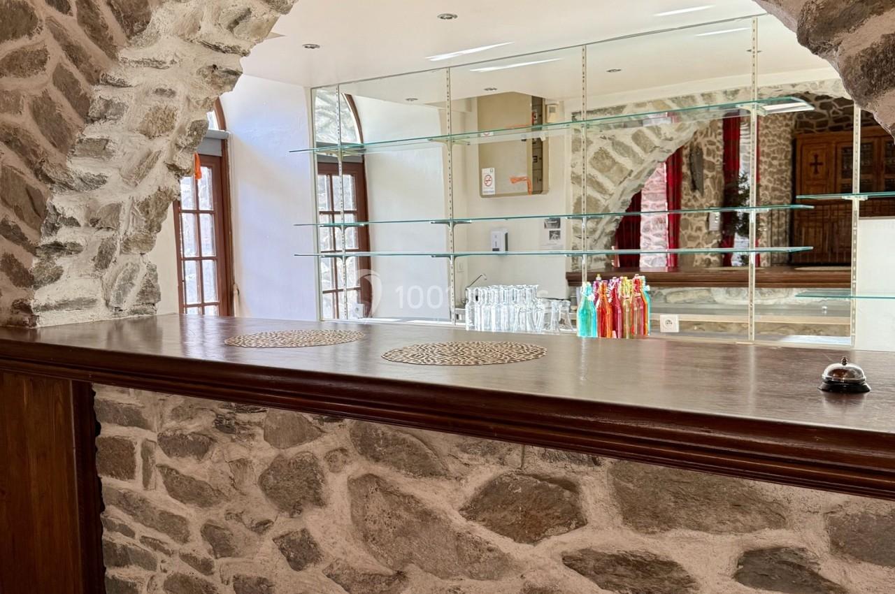 Comptoir en bois devant un mur en pierre, avec étagères en verre, bouteilles colorées et lumière naturelle.