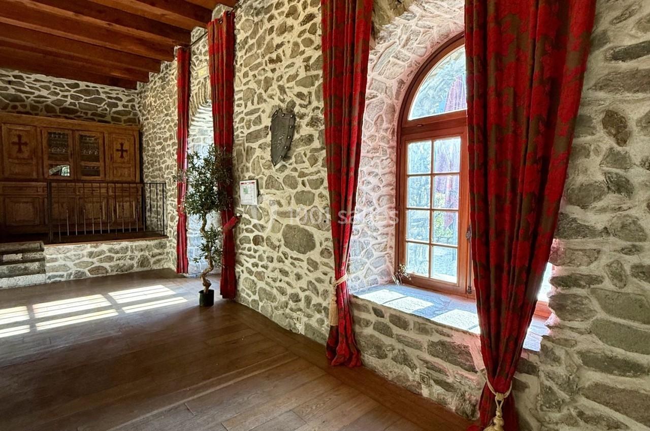 Mur en pierre avec une grande fenêtre cintrée, encadrée de rideaux rouges, laissant entrer la lumière naturelle.