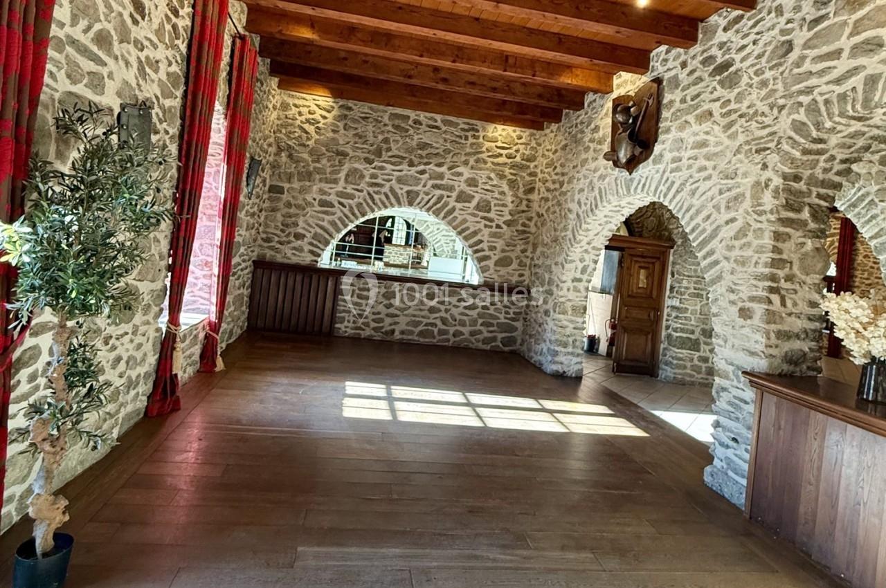 Salle en pierre avec poutres apparentes, grandes arches, parquet en bois et lumière naturelle entrant par les fenêtres.