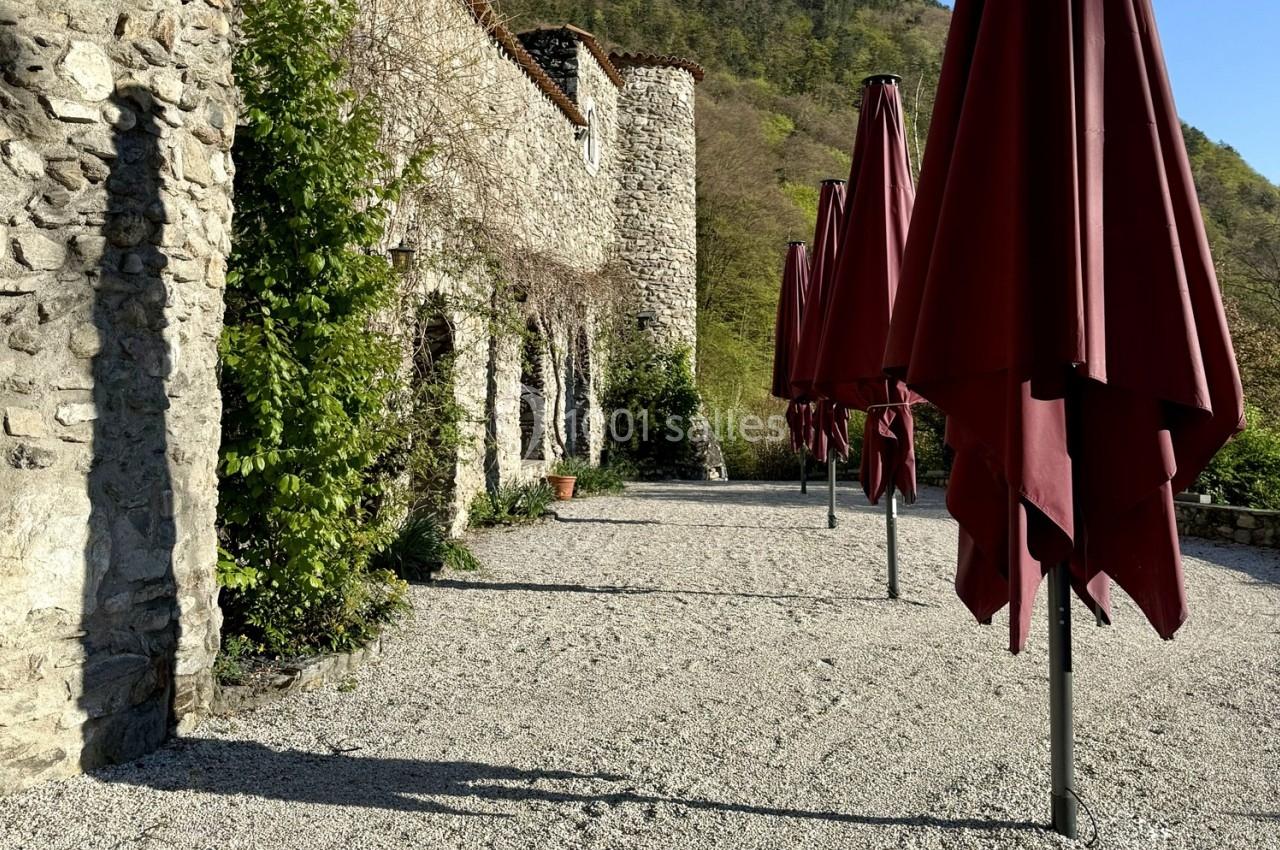 Terrasse en gravier avec parasols rouges fermés, bordée par un bâtiment en pierre et une végétation verdoyante.