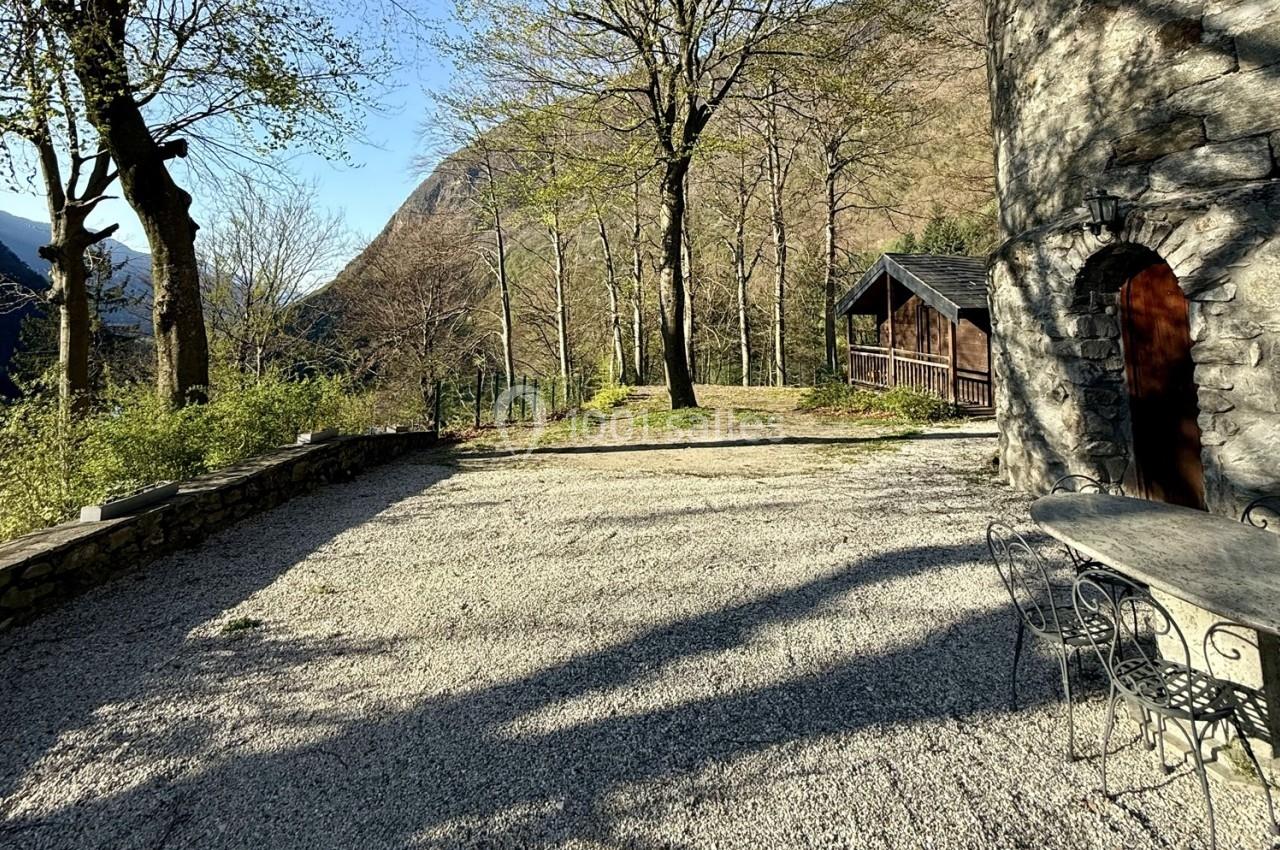 Cour en gravier entourée d'arbres, avec une maison en pierre et une table en métal à droite, vue sur des collines boisées.