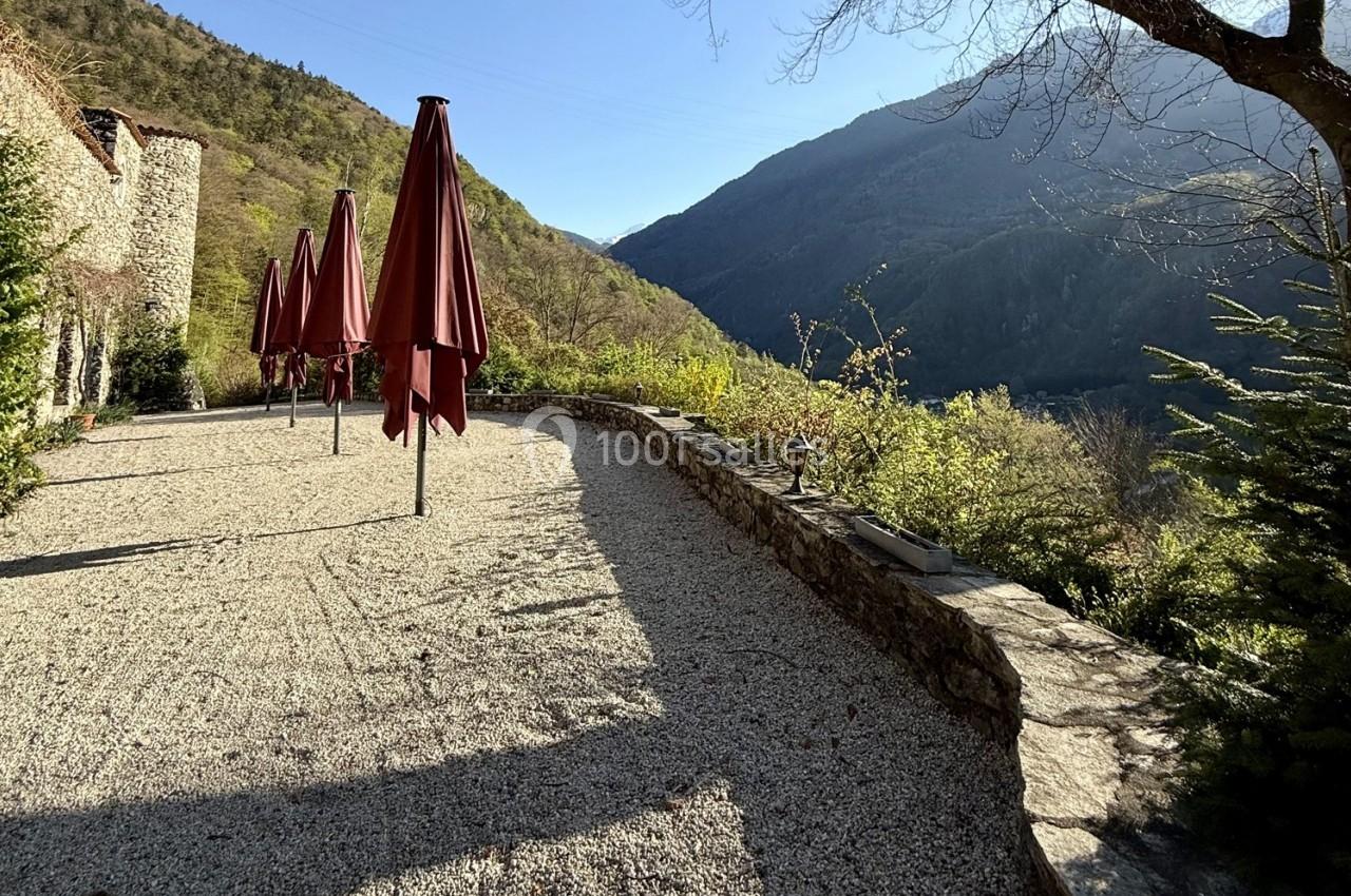 Terrasse en gravier avec parasols rouges, bordée d'un muret en pierre, offrant une vue sur des montagnes verdoyantes.