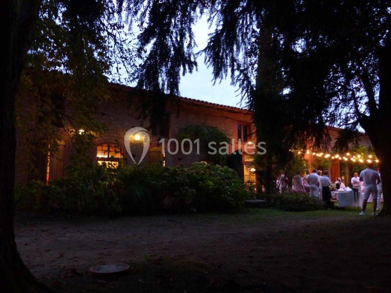 Bâtiment en briques éclairé à la tombée de la nuit, entouré d'arbres, avec des convives rassemblés à l'extérieur.