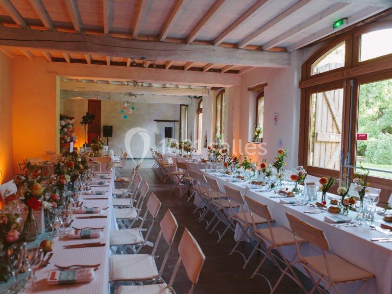 Salle de réception lumineuse avec de longues tables décorées de fleurs, chaises blanches et grandes fenêtres ouvertes sur l…
