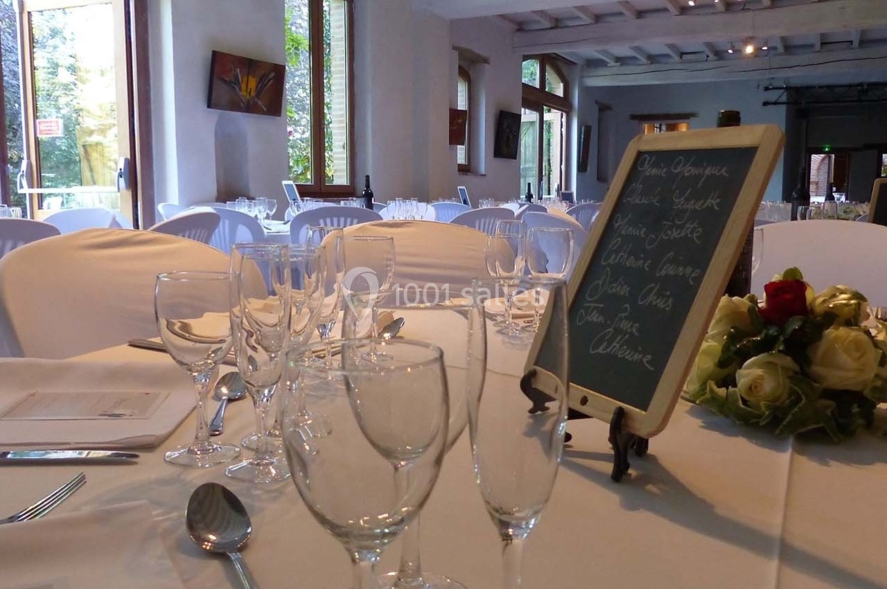 Salle de réception lumineuse avec tables dressées, nappes blanches, verres, couverts et ardoise de menu.