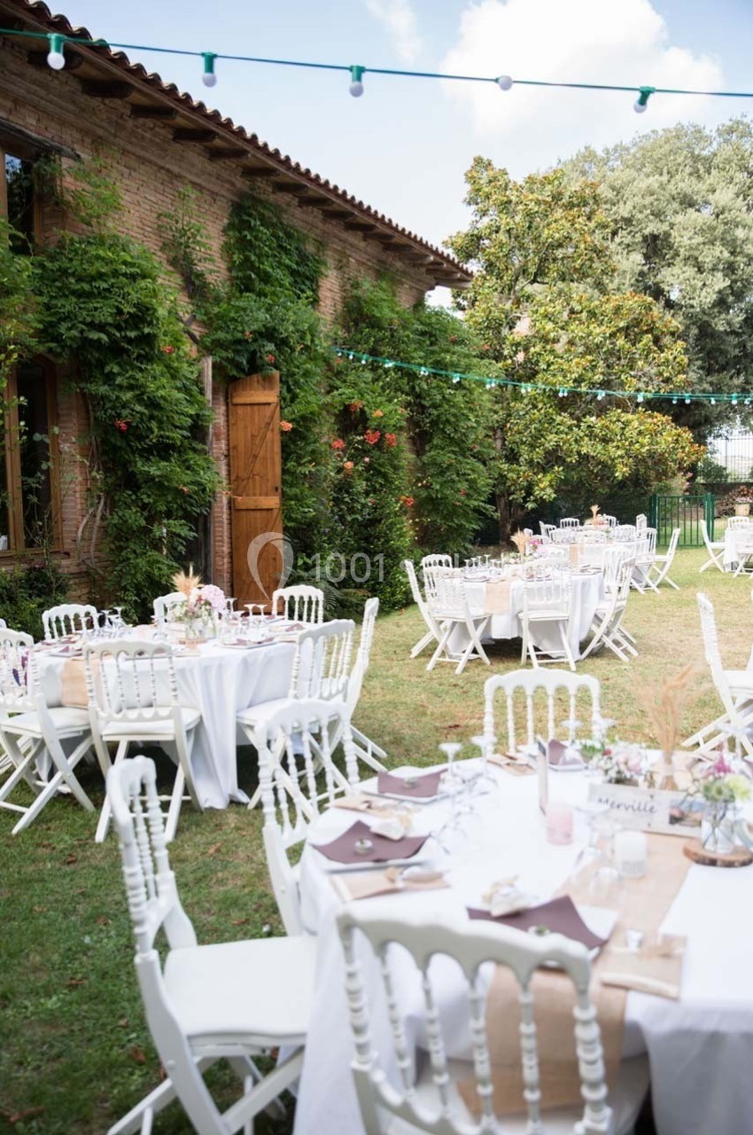 Tables rondes décorées pour un événement en plein air, disposées dans un jardin avec une façade en briques et des guirlandes…