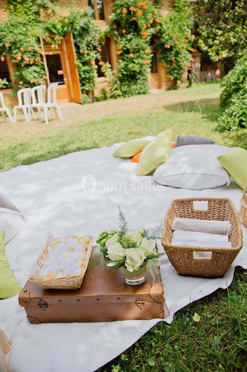 Tapis blanc sur pelouse avec coussins colorés, paniers en osier, fleurs et table basse en bois dans un jardin verdoyant.