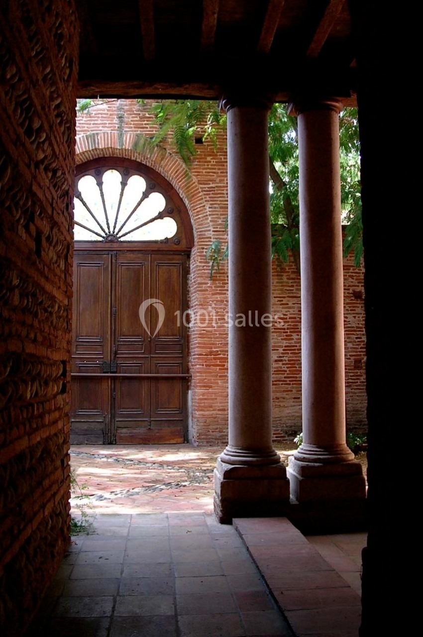 Vue d'une porte en bois encadrée par des colonnes et un mur de briques, éclairée par la lumière extérieure.