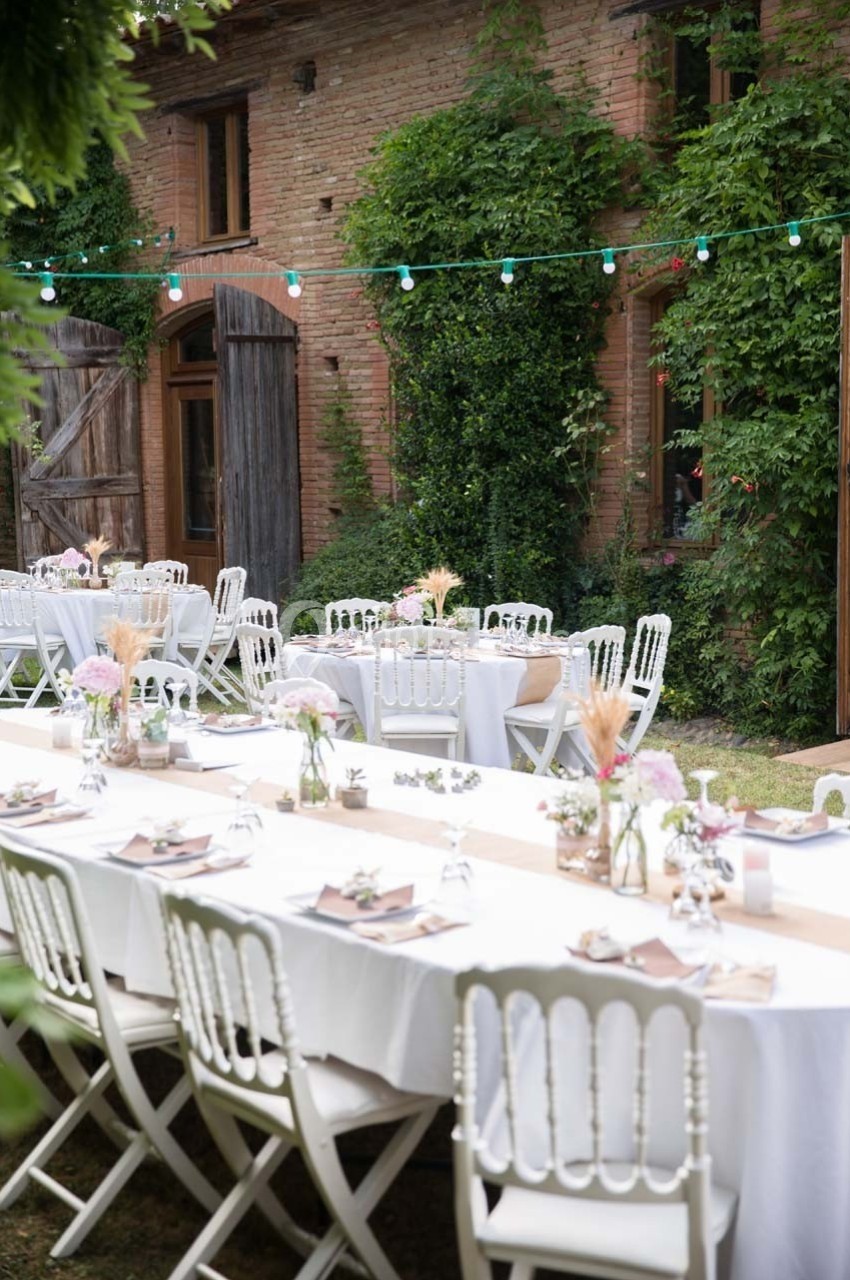 Tables décorées pour un événement en extérieur, disposées devant un bâtiment en briques avec des guirlandes lumineuses.