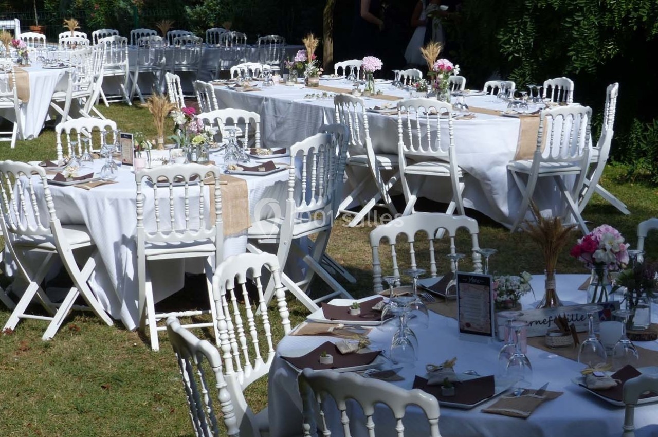 Tables de réception décorées avec nappes blanches, fleurs et vaisselle, disposées dans un jardin en plein air.