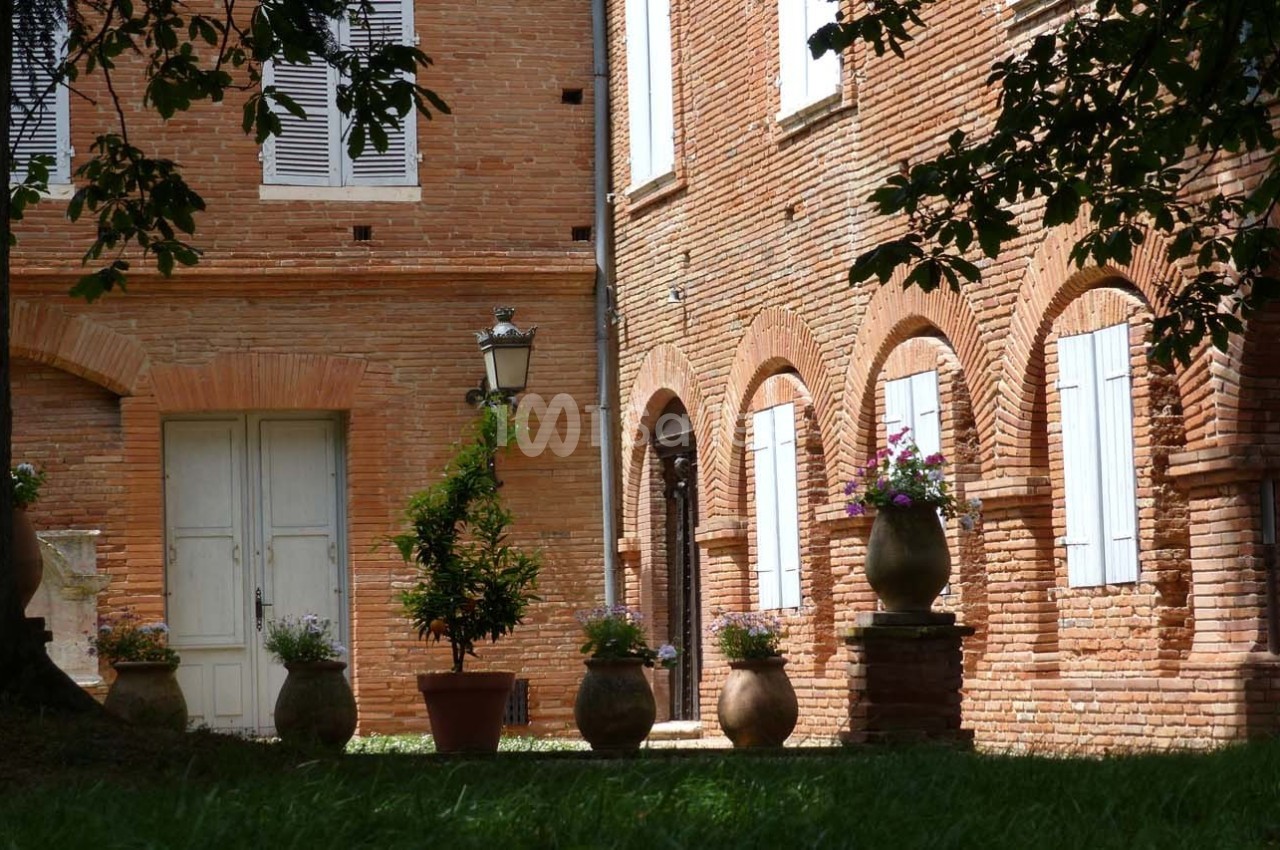 Cour bordée de murs en briques rouges avec des arches, des fenêtres à volets blancs et des pots de fleurs alignés.