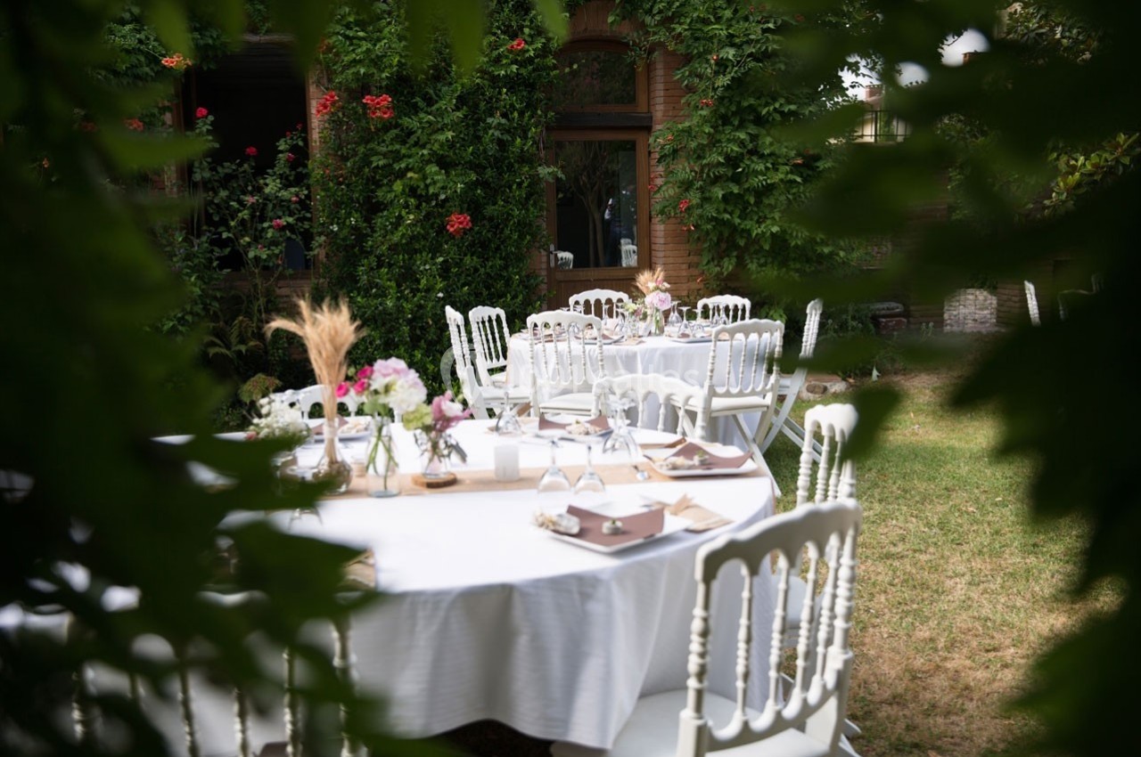 Tables rondes dressées avec nappes blanches et décorations florales dans un jardin verdoyant.