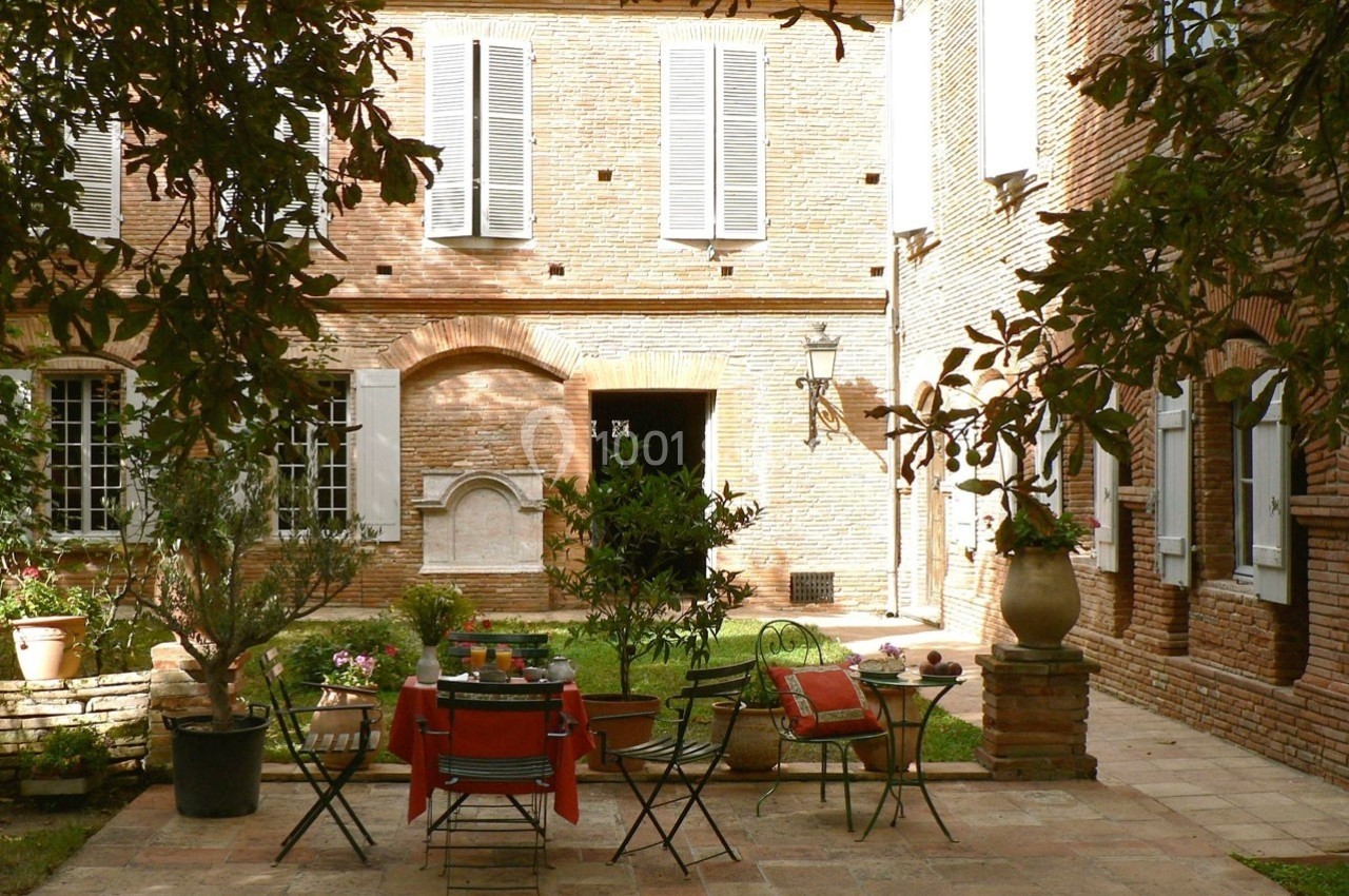 Cour intérieure avec des tables et chaises en métal, entourées de plantes, devant une maison en briques avec volets blancs.