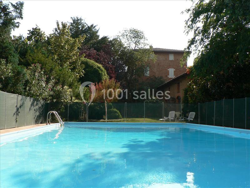 Piscine extérieure entourée de verdure avec une maison en briques en arrière-plan par une journée ensoleillée.