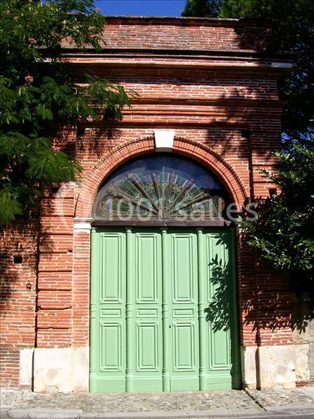 Porte en bois vert clair avec un arc en brique rouge et un vitrail en éventail, entourée de végétation.