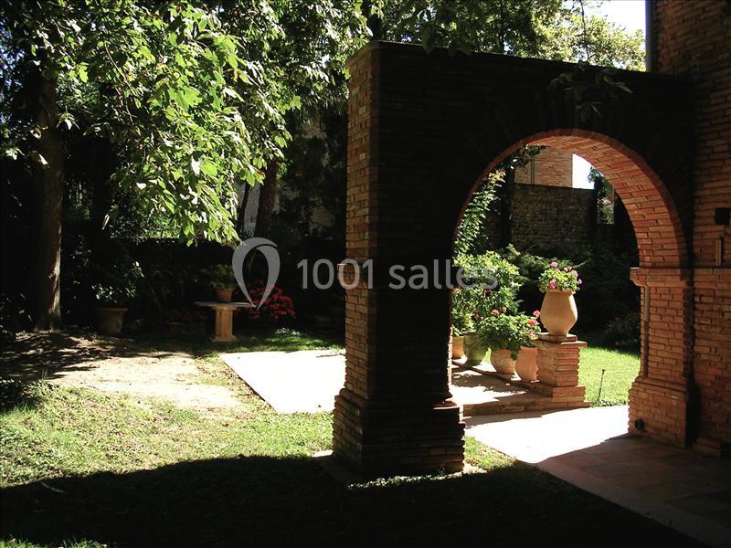 Arc en briques dans un jardin ombragé, entouré de végétation et de pots de fleurs sur des socles.