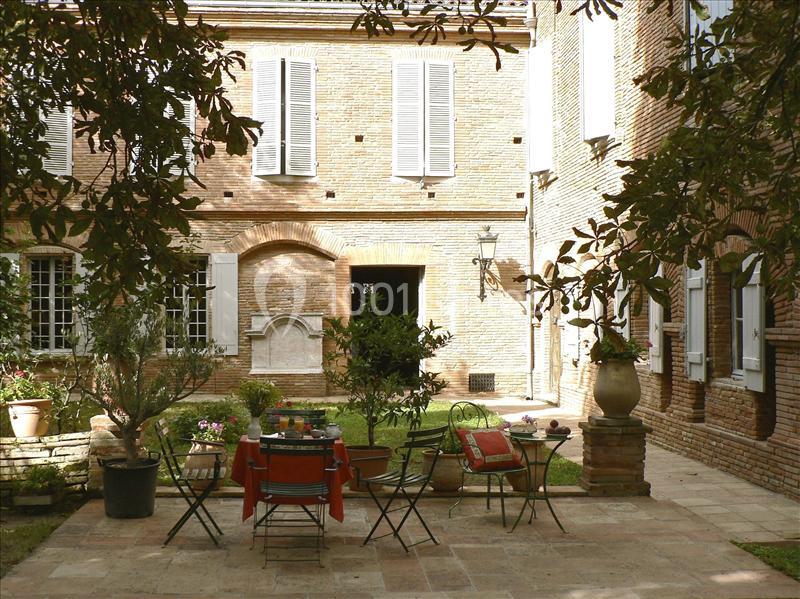 Cour intérieure avec table et chaises en métal, entourée de végétation, devant une façade en briques avec volets blancs.