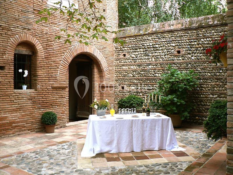 Table dressée avec nappage blanc dans une cour en briques et galets, entourée de plantes et bougies décoratives.