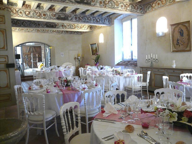 Salle de réception décorée avec des tables rondes dressées, nappes blanches et touches de rose, sous un plafond orné.