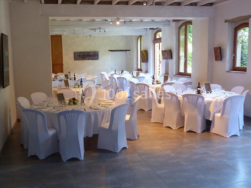 Salle de réception lumineuse avec tables rondes dressées, nappes blanches et chaises couvertes, prête pour un événement.