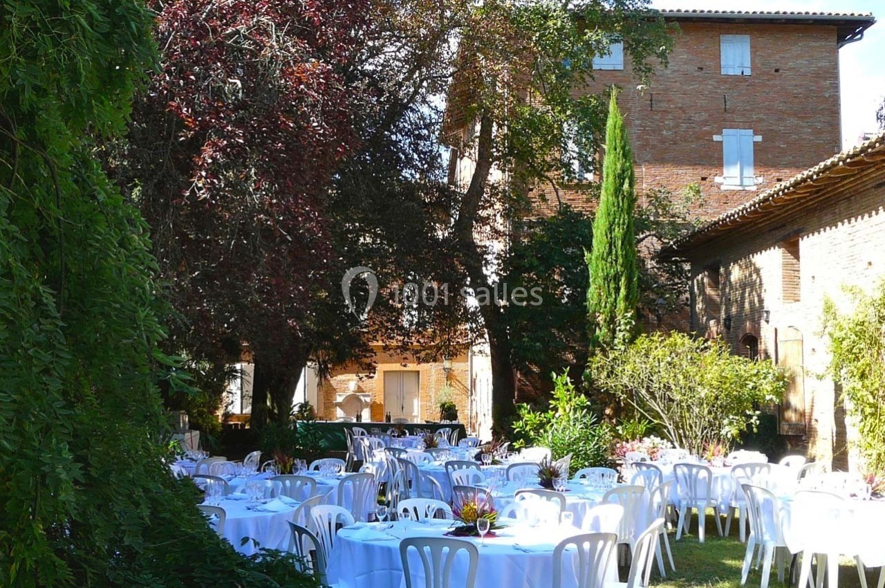 Tables rondes dressées avec nappes blanches dans un jardin arboré, devant un bâtiment en briques.