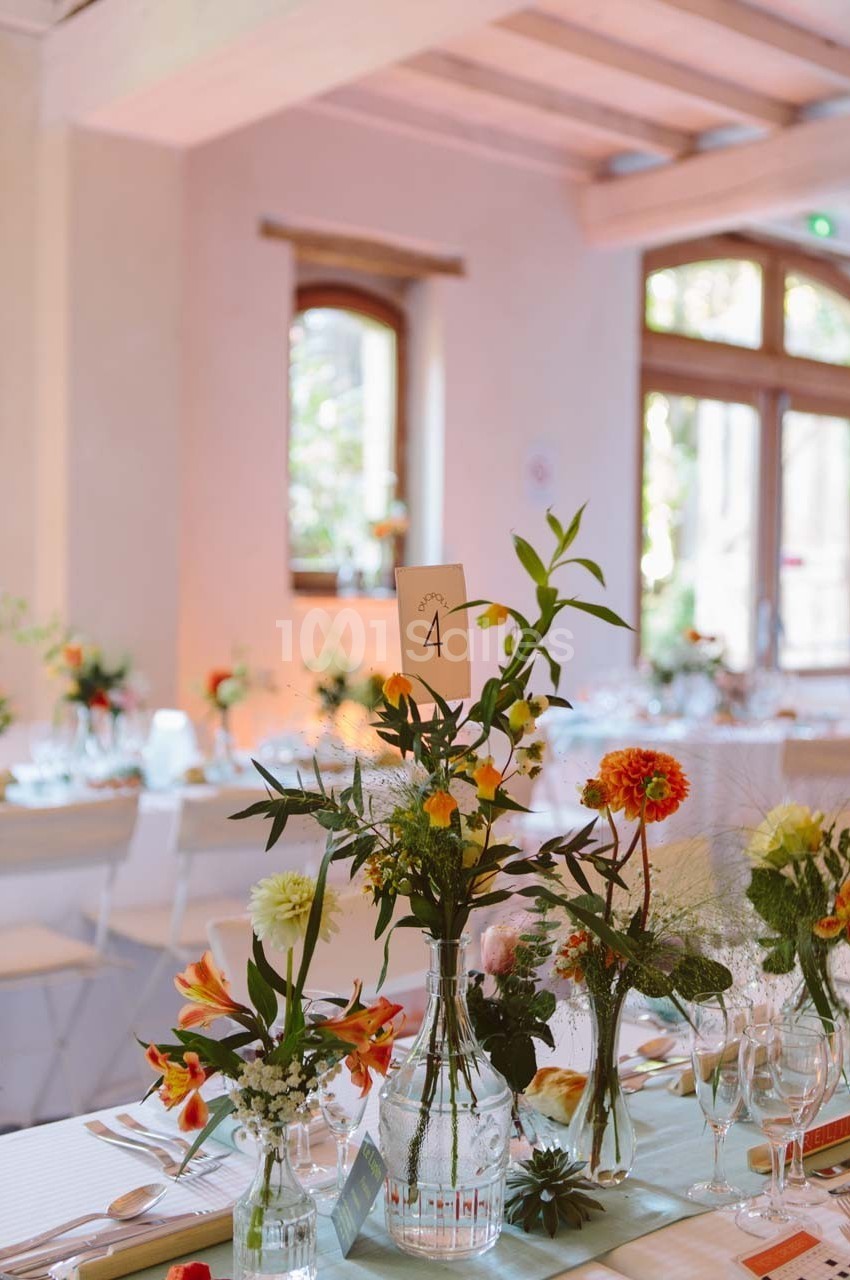 Décoration de table avec des fleurs colorées et un numéro de table ’4’ dans une salle lumineuse aux grandes fenêtres.