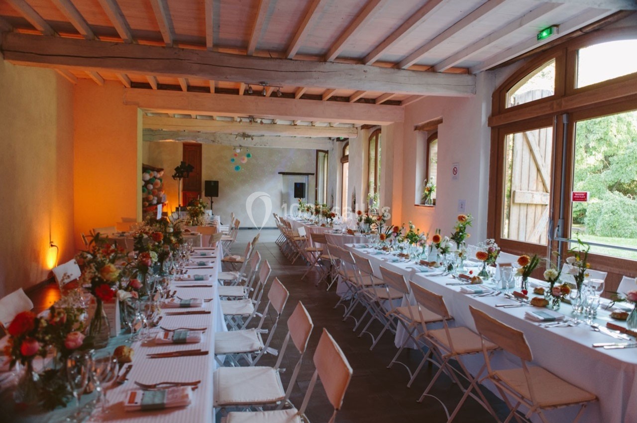 Salle de réception décorée avec des tables alignées, nappes blanches, chaises pliantes et arrangements floraux colorés.