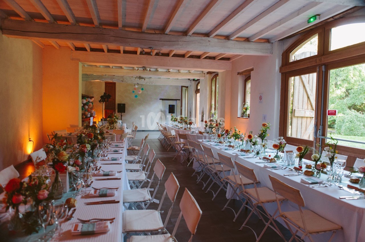 Salle de réception décorée avec des tables alignées, nappes blanches, chaises pliantes et arrangements floraux colorés.