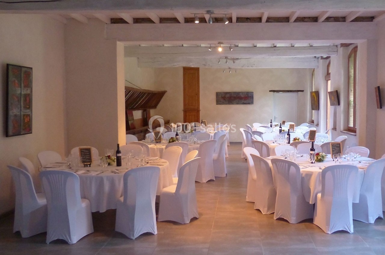 Salle de réception lumineuse avec tables rondes dressées, nappes blanches et chaises couvertes, prête pour un événement.