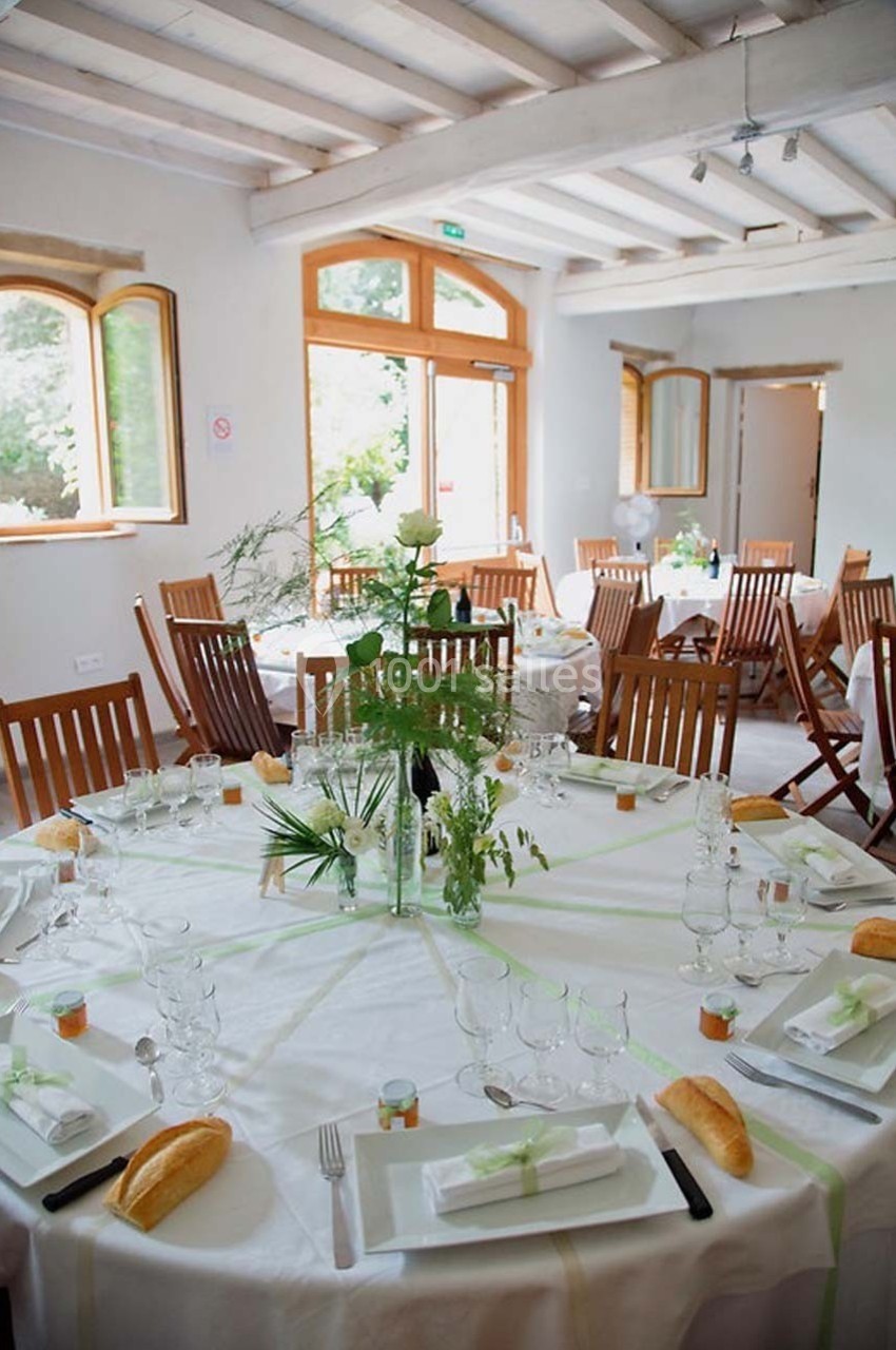 Salle lumineuse avec tables rondes dressées pour un repas, décorées de nappes blanches, plantes et vaisselle élégante.
