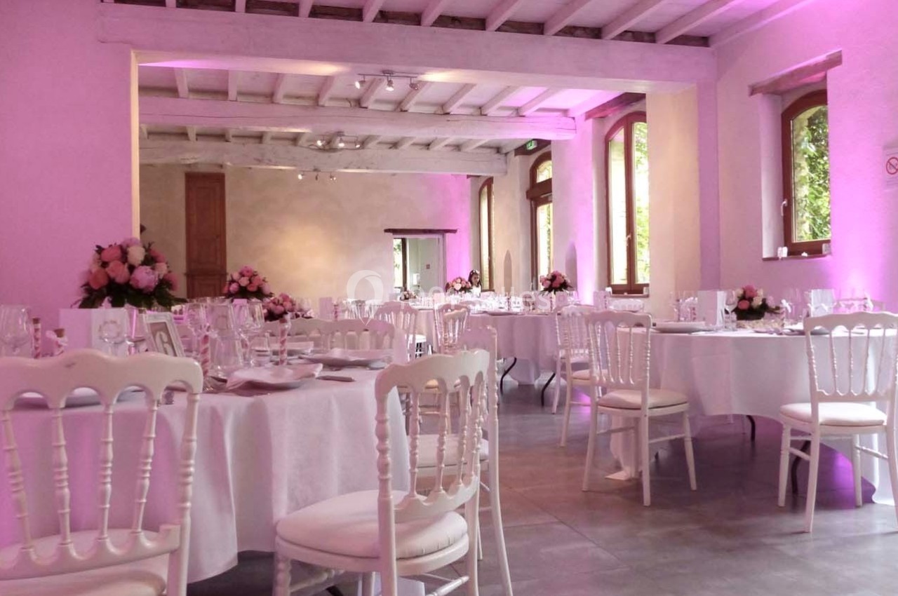 Salle de réception décorée avec des tables rondes dressées, chaises blanches et éclairage rose.