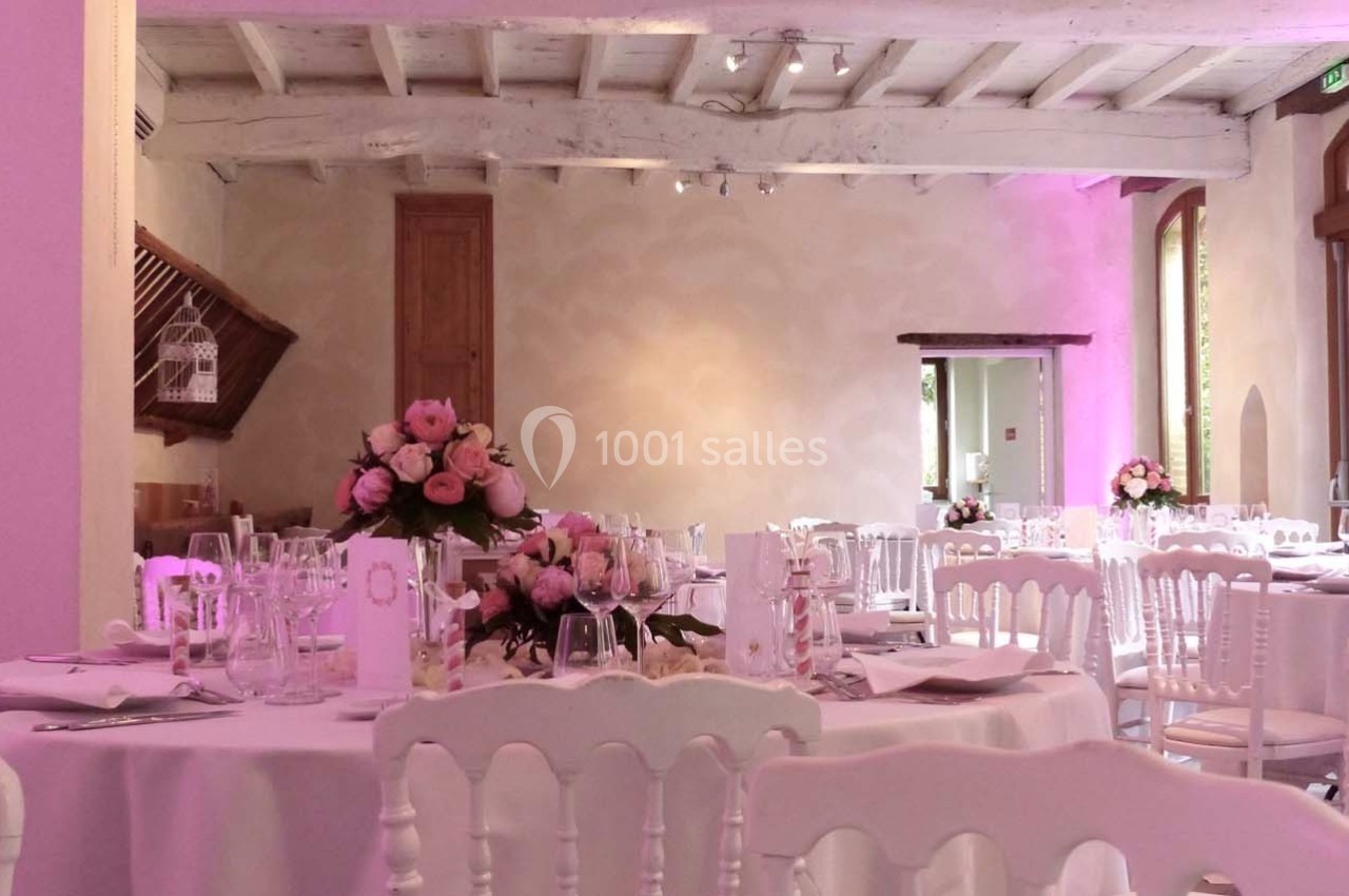 Salle de réception décorée avec des tables rondes, nappes blanches, chaises assorties et bouquets de fleurs roses.