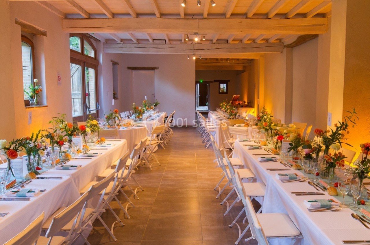 Salle décorée pour un événement, avec deux longues tables dressées, chaises blanches et arrangements floraux colorés.