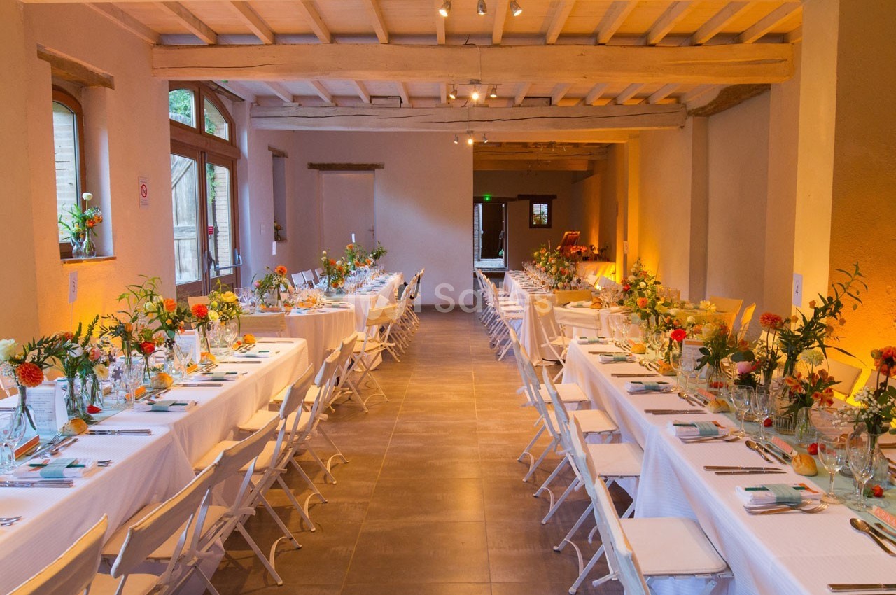 Salle décorée pour un événement, avec deux longues tables dressées, chaises blanches et arrangements floraux colorés.