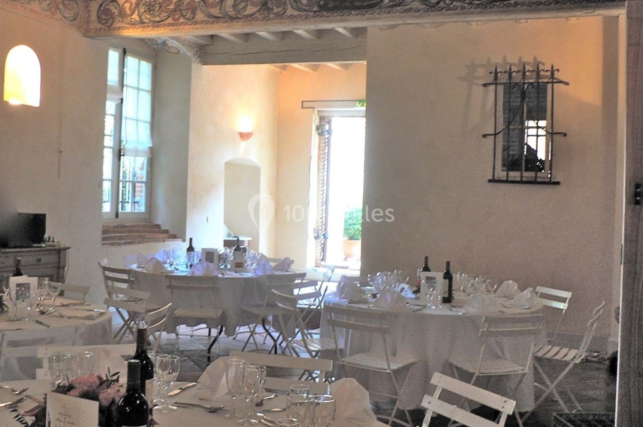 Salle de réception lumineuse avec tables dressées, chaises blanches pliantes et décorations murales en pierre.