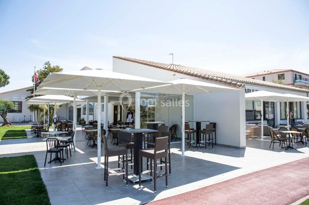 Terrasse extérieure avec tables, chaises et parasols, entourée de pelouse et bâtiments modernes en arrière-plan.