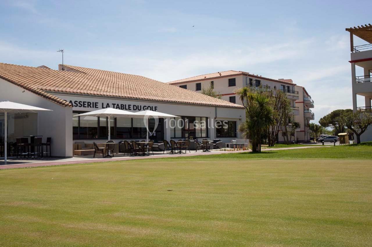Terrasse d'une brasserie avec parasols, située près d'un terrain de golf et entourée de bâtiments modernes.