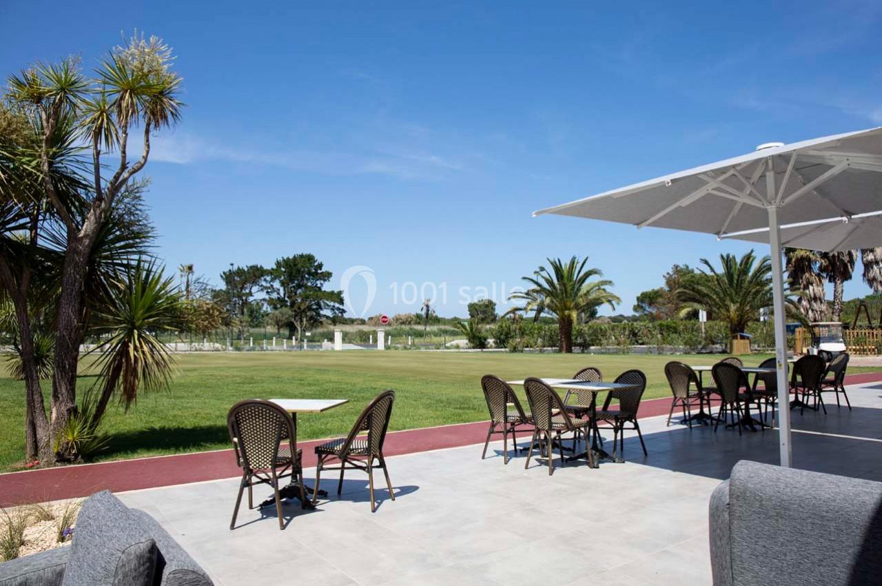 Terrasse extérieure avec tables et chaises, vue sur une pelouse, palmiers et un ciel bleu dégagé.