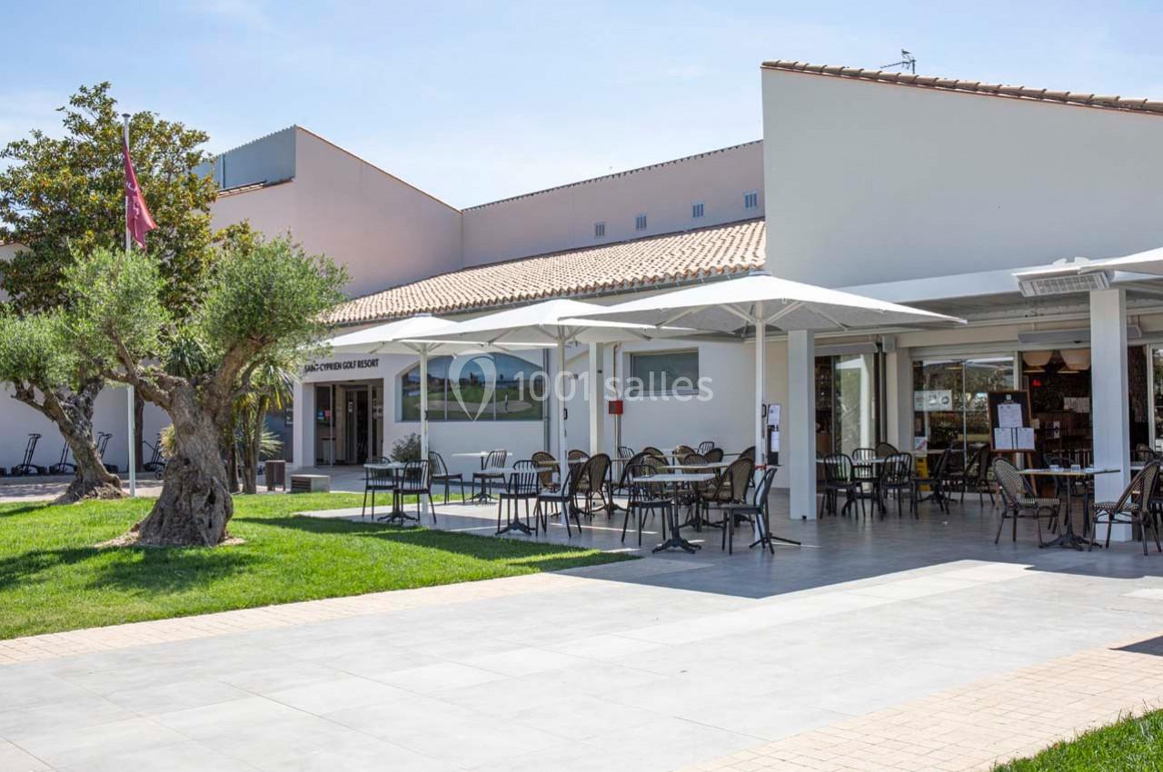 Terrasse extérieure d'un bâtiment moderne avec tables, chaises, parasols et un olivier sur une pelouse.