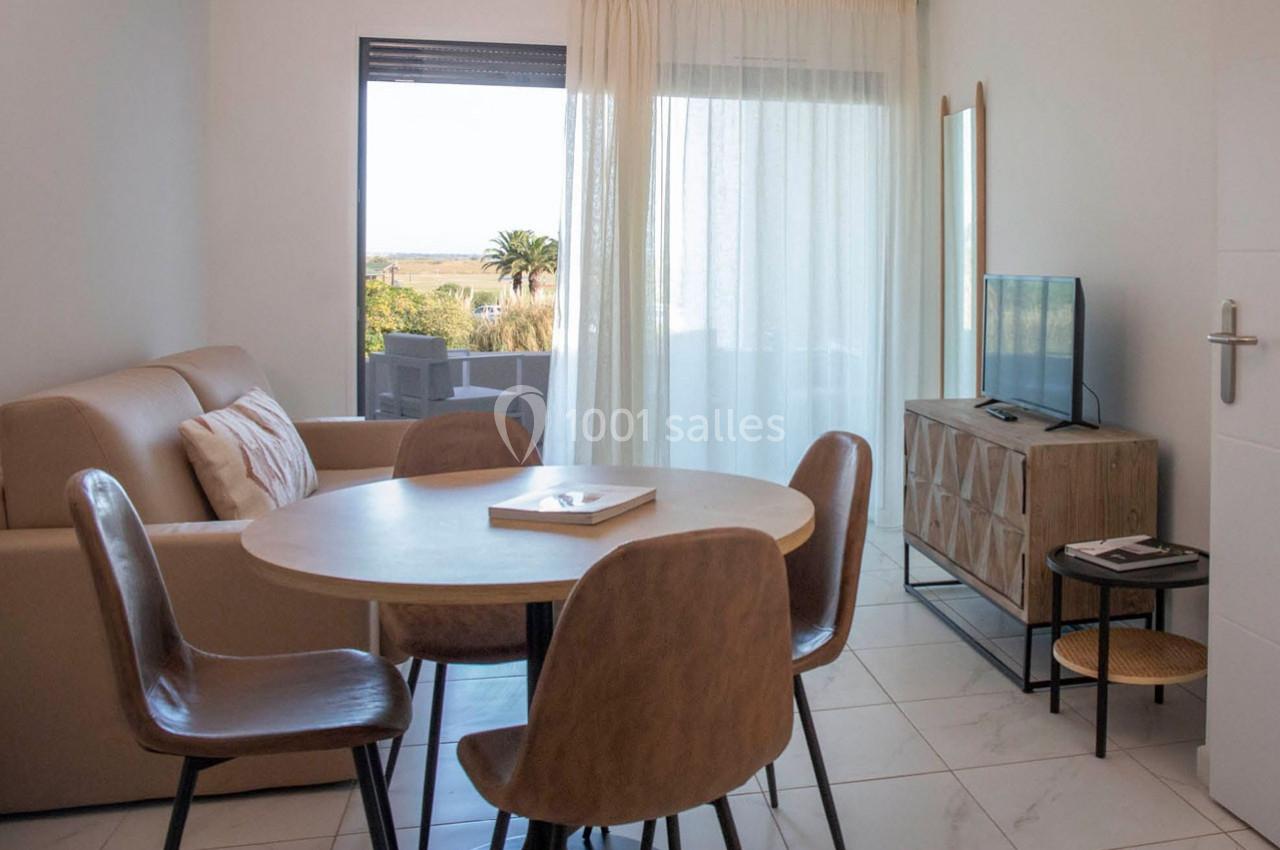 Salon lumineux avec table ronde, chaises en bois, canapé beige, meuble TV et grande baie vitrée donnant sur l'extérieur.