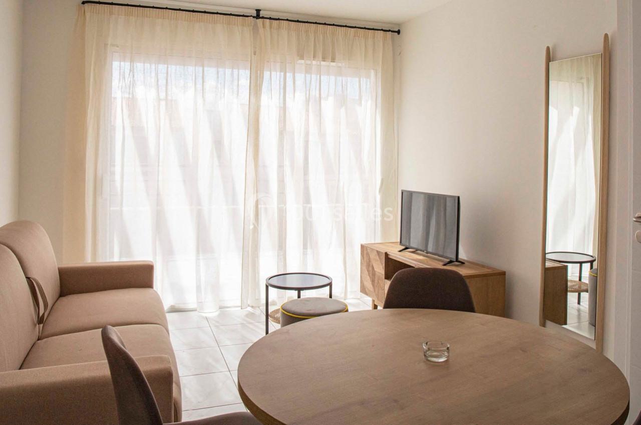 Salon lumineux avec canapé beige, table ronde en bois, télévision sur meuble et grande baie vitrée avec rideaux clairs.