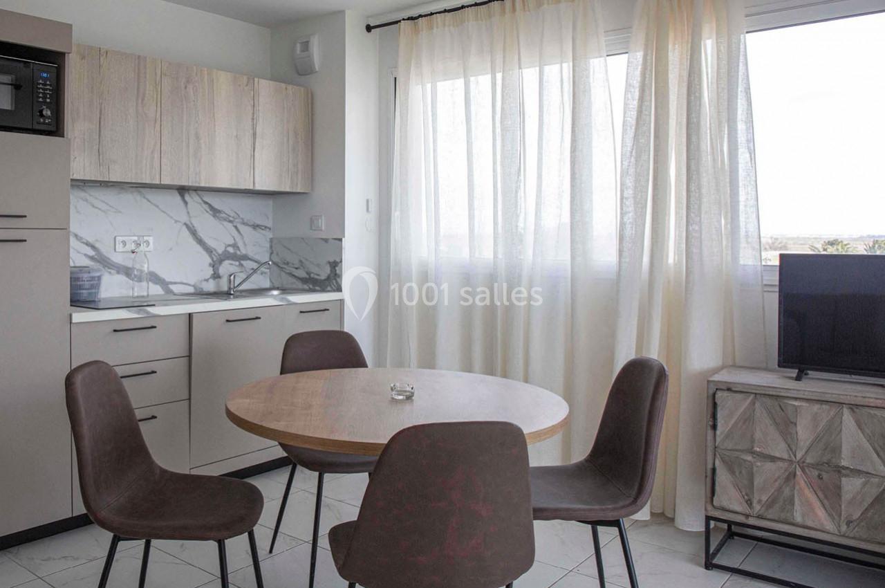 Cuisine ouverte avec table ronde en bois, quatre chaises, un meuble TV, et une grande fenêtre avec rideaux clairs.