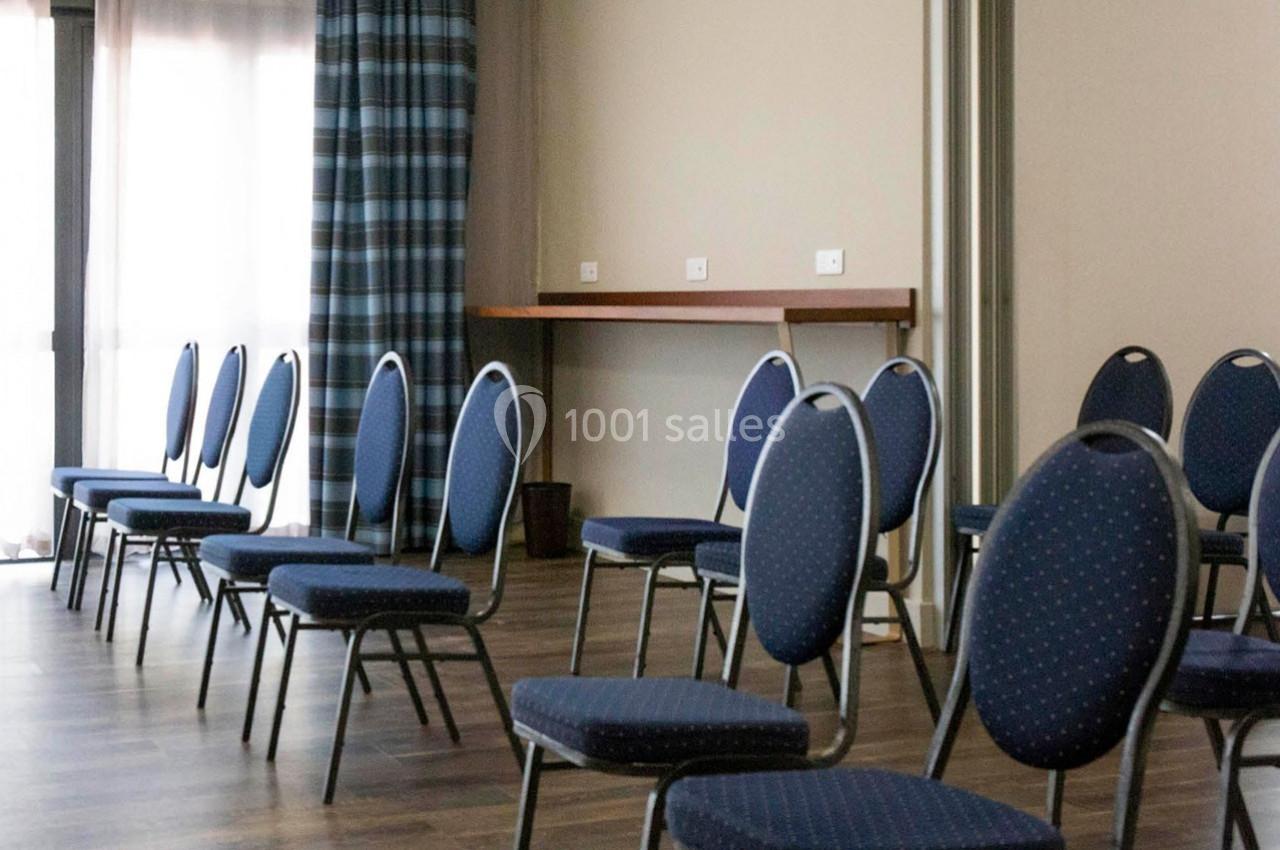 Chaises bleues disposées en rangées dans une salle lumineuse avec parquet et rideaux à carreaux.