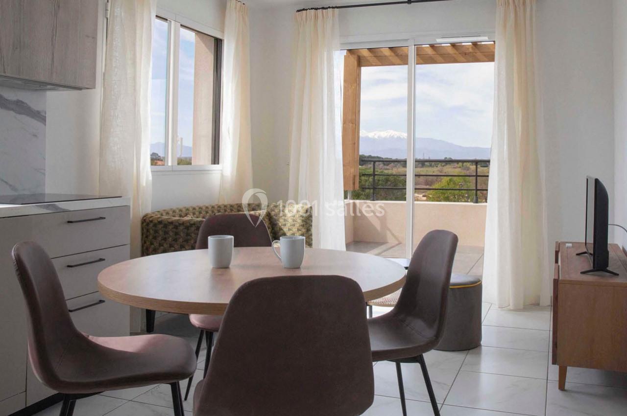 Salle à manger lumineuse avec table ronde, chaises, canapé, télévision et vue sur une terrasse avec paysage montagneux.