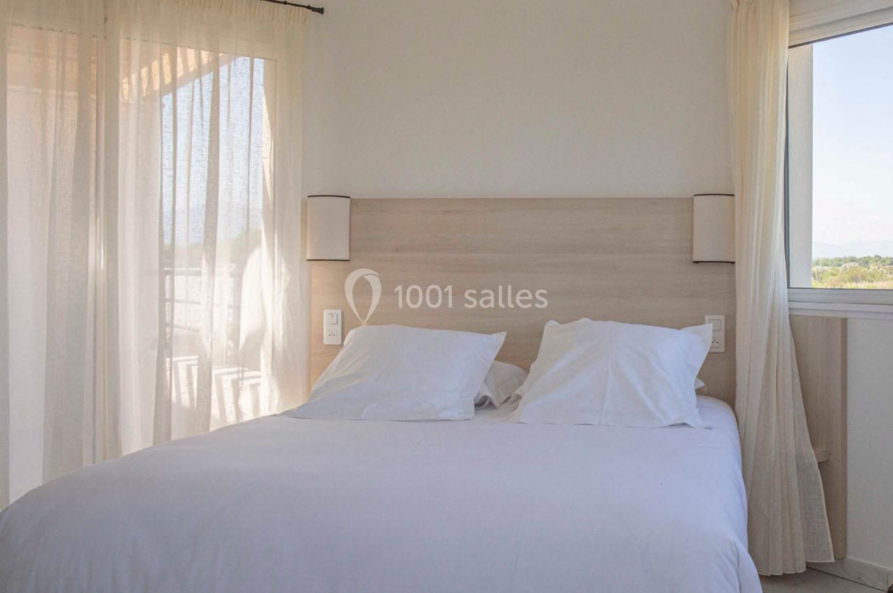 Lit double avec draps blancs dans une chambre lumineuse, encadrée par des rideaux clairs et une fenêtre donnant sur l…