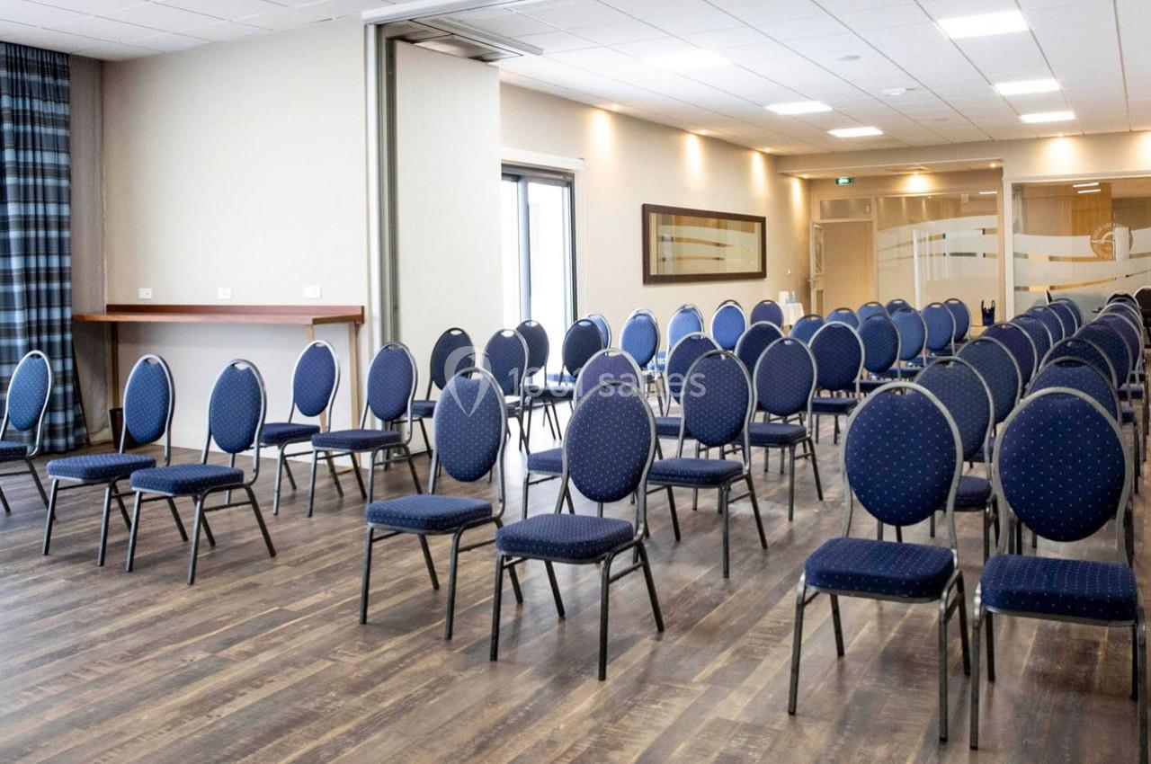 Salle de réunion avec des chaises bleues disposées en rangées sur un sol en bois, éclairée par des lumières au plafond.