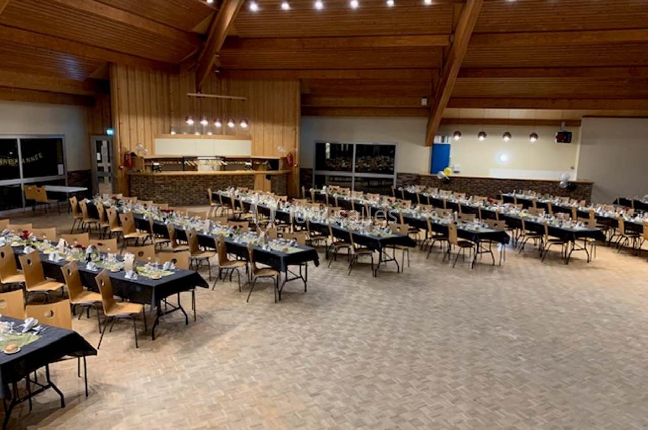 Salle spacieuse avec des tables rectangulaires dressées pour un événement, éclairée par des lumières suspendues.